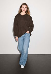 Brun oversized tröja med V-ringning, ihop med ljusblå vida jeans. Modellen bär vita sneakers och står mot en neutral bakgrund.