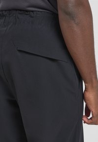 Schwarze Hosen mit lockerer Passform, mit einem elastischen Bund und einer Klappentasche auf der rechten Seite in der Nähe der Hüfte. Glatte Textur.