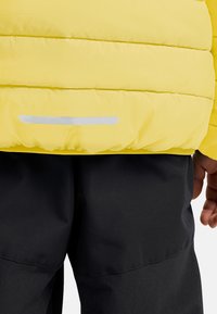 Chaqueta acolchada amarilla con costuras horizontales, que presenta una cinta reflectante en el dobladillo, combinada con pantalones negros. La tela parece suave.