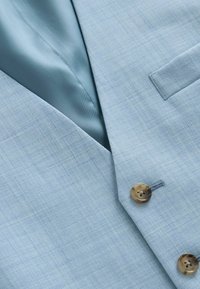 Veste de costume bleu clair avec un revers cranté, des boutons marron et une doublure intérieure lisse assortie visible près du col.