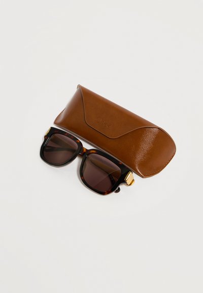 Τετράγωνα γυαλιά ηλίου με μοτίβο χελώνας, σκοτεινούς φακούς και χρυσές λεπτομέρειες δίπλα σε καφέ δερμάτινη θήκη γυαλιών Chloé σε ανοιχτό φόντο.