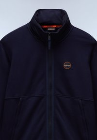 Marineblå zip-up jakke laget av et mykt stoff med høy krage, med to frontlommer og et rundt logo på venstre bryst.