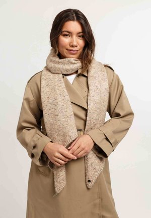 Donna con un trench beige e una sciarpa spessa beige, in piedi con le mani delicatamente intrecciate davanti a uno sfondo chiaro e semplice.