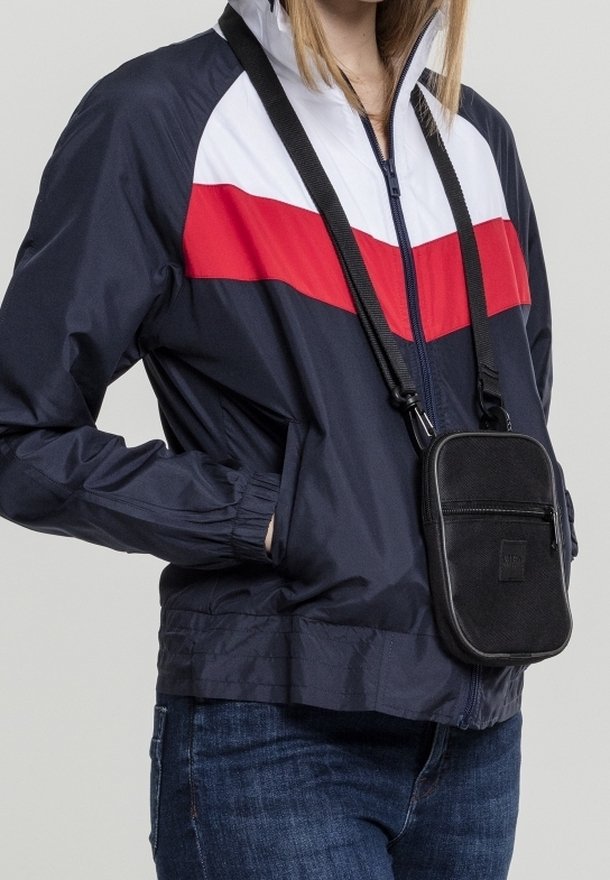 Marinblå vindjacka med vita och röda horisontella ränder, elastiska ärmslut, i kombination med en liten svart crossbody-väska. Slät textur.