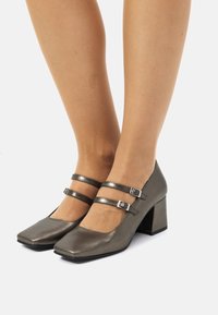 Zapatos de tacón Mary Jane metálicos grises con puntas cuadradas, dos correas con hebillas y tacones cuadrados. Textura suave con un ligero brillo.