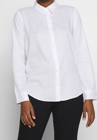 Weißes, langärmliges Hemd mit Knopfleiste und Kragen, mit einer glatten Textur und abgerundetem Saum, kombiniert mit schwarzen Hosen.