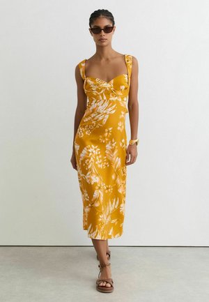 Vrouw draagt een mosterdgele midi-jurk met witte bloemenprint, bruine sandalen, een zonnebril en een armband, staand tegen een witte muur.
