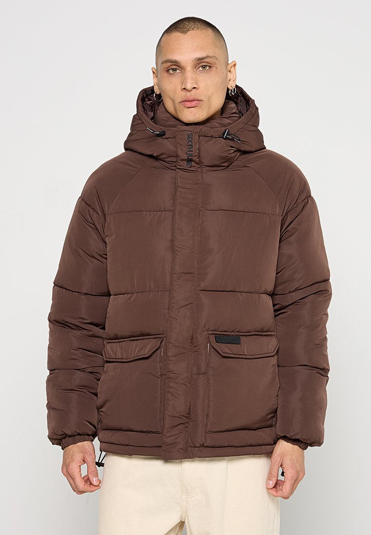 Mann in einer braunen gepolsterten Winterjacke mit Kapuze und zwei aufgesetzten Taschen, der vor einem schlichten hellen Hintergrund steht.