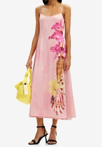 Mujer vestida con un vestido midi rosa con estampado de flores y conchas marinas, sosteniendo un bolso amarillo, acompañado de sandalias de tacón negro.
