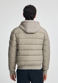 Giubbotto imbottito color beige chiaro con imbottitura trapuntata, cappuccio aderente e polsini a costine. Presenta una texture di tessuto liscia e una vestibilità morbida.
