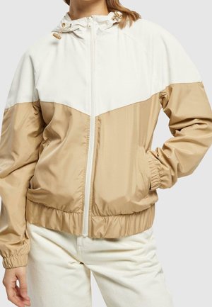 Femme portant une veste à capuche beige et blanc cassé zippée, avec poignets et taille élastiques, debout avec une main dans une poche.