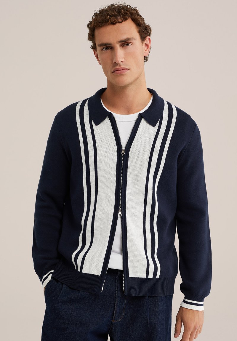 Homme aux cheveux bouclés portant un cardigan zippé bleu marine et blanc à rayures verticales sur une chemise blanche et un pantalon foncé.