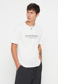 Λευκό βαμβακερό μπλουζάκι με κοντά μανίκια και μαύρο τυπωμένο λογότυπο. Συνδυασμένο με αέρινες σκούρες γκρι παντελόνες, παρουσιάζοντας έναν μινιμαλιστικό σχεδιασμό.