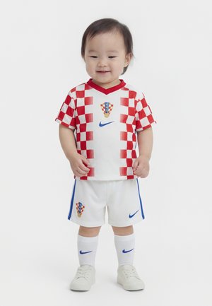 Tout-petit portant un maillot de football à carreaux rouges et blancs, un short blanc avec des rayures bleues, des chaussettes blanches et des baskets blanches, souriant et debout.
