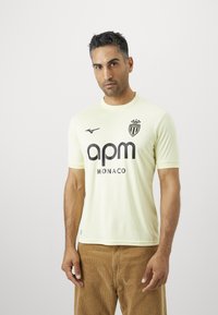 Svetložltý futbalový dres s krátkymi rukávmi, ktorý má okrúhly výstrih, textúrovanú tkaninu a čierne logo s textom "apm MONACO" na prednej strane.