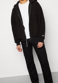 Sudadera de forro polar negra con cremallera, capucha, bolsillos frontales y detalle de cremallera en contraste. Combinada con vaqueros negros de pierna recta. Textura suave.