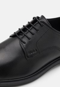 Chaussure en cuir noir avec dessus texturé, design traditionnel à lacets, semelle en caoutchouc et marquage discret sur le côté.