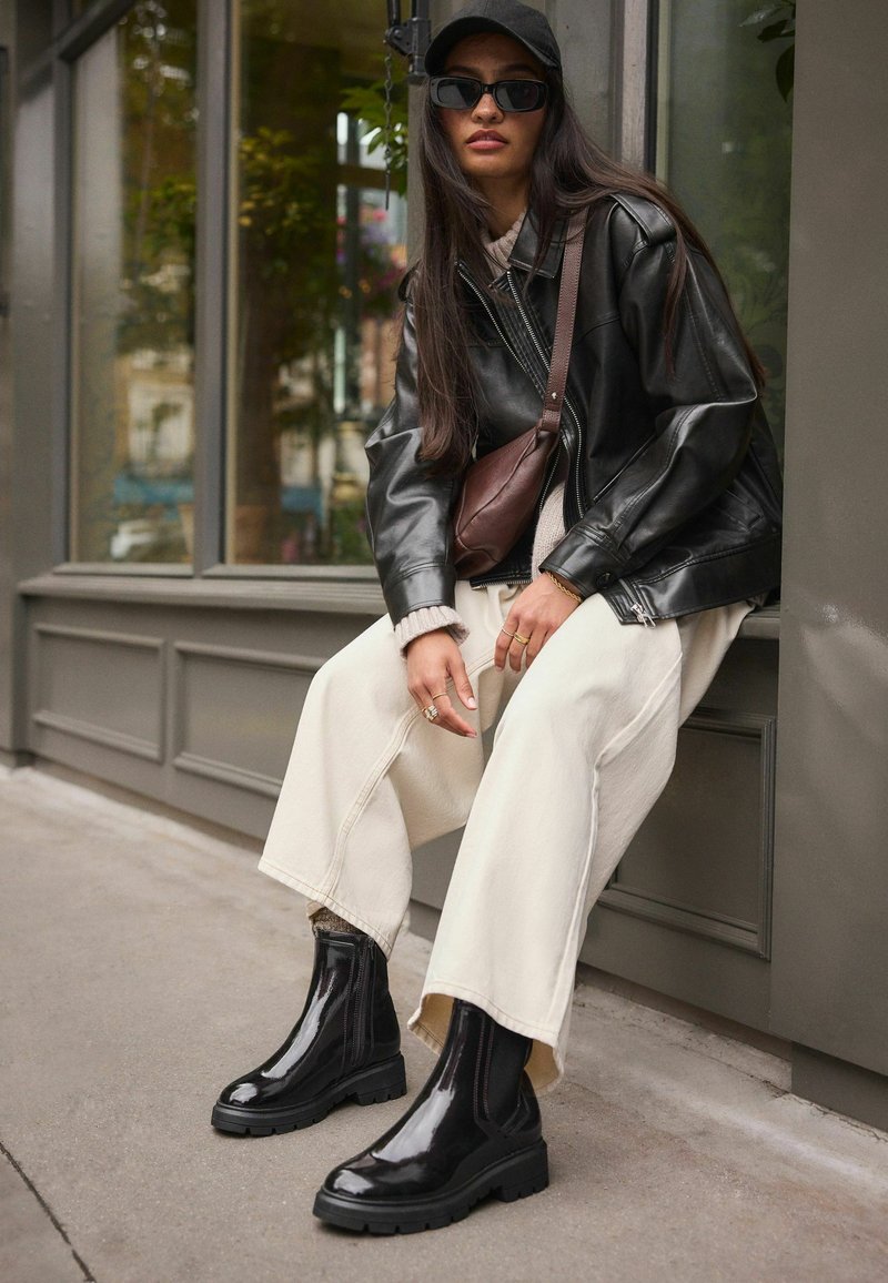 Schwarze Lederjacke, cremiger Pullover, weite cremige Hose, glänzende schwarze Stiefeletten, braune Umhängetasche, langes dunkles Haar, Sonnenbrille und Mütze.