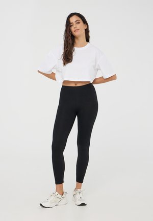 T-shirt bianco a maniche corte, tagliato corto; abbinato a leggings neri. La modella indossa scarpe sportive bianche con accenti grigi. Design semplice, senza motivi.