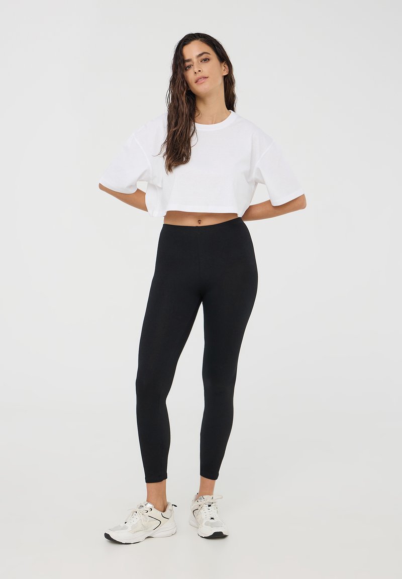 T-shirt bianco a maniche corte, tagliato corto; abbinato a leggings neri. La modella indossa scarpe sportive bianche con accenti grigi. Design semplice, senza motivi.