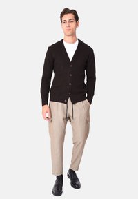 Uomo con cardigan nero abbottonato, t-shirt bianca, pantaloni cargo beige e mocassini neri in piedi con le mani nelle tasche.