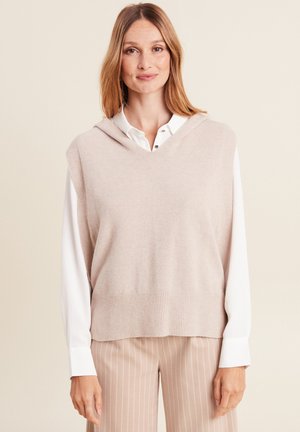 Gilet en tricot rose clair avec capuche, présentant un design en V et un ourlet côtelé. Porté par-dessus une chemise blanche à col. Pantalons beiges à rayures.
