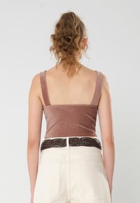 Braunes Cord-Crop-Top mit breiten Trägern, kombiniert mit hochtaillierten cremefarbenen Hosen und einem geflochtenen braunen Gürtel. Glatte Textur, taillierte Passform.