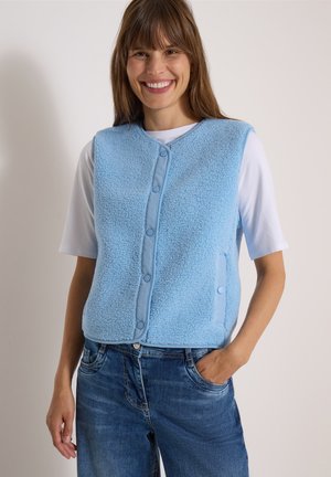 Smilende kvinde iført en lyseblå fleecervest med knapper over en hvid kortærmet skjorte og blå jeans med den ene hånd i lommen.