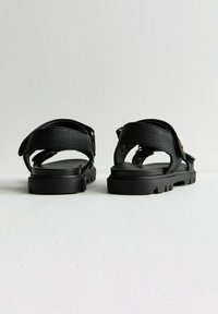 Svarta sandaler gjorda av texturerat material med justerbara remmar. Sulan är tjock och robust, med en platt design och utskurna detaljer.
