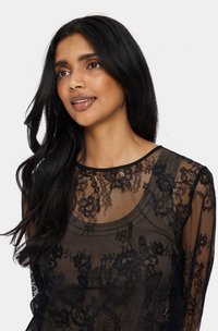 Fekete csipke felső hosszú ujjakkal, virágmintákkal, kerek nyakkivágással és áttetsző textúrával. Könnyű és testhezálló kialakítás.