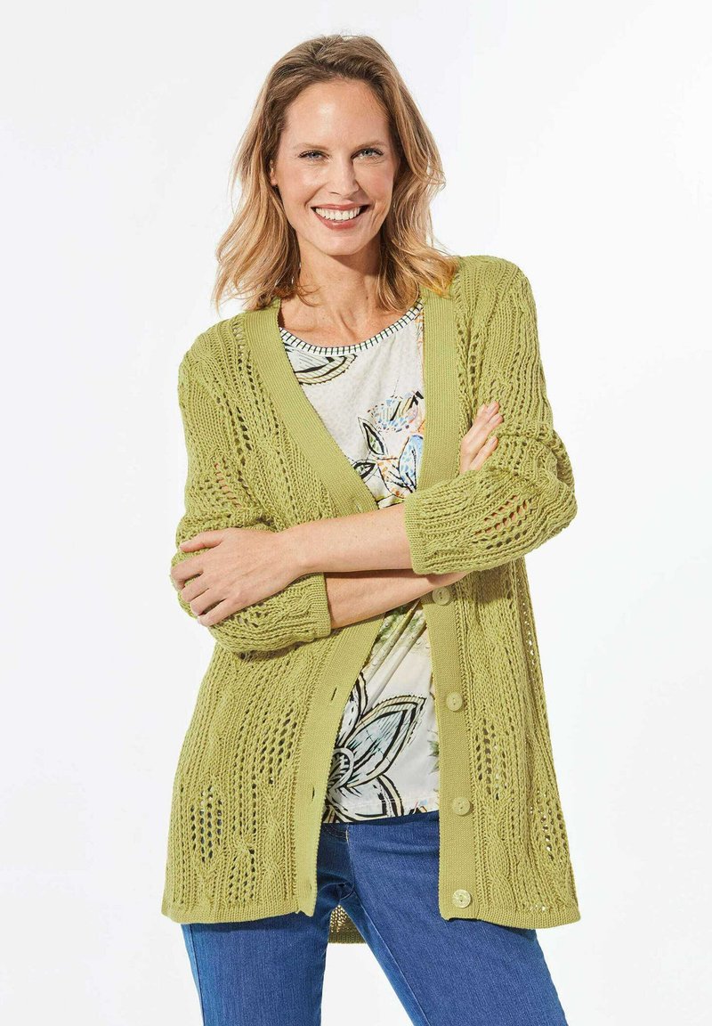 Hellgrüner, gestrickter Cardigan mit einem lockeren, offenen Design, das strukturierte Muster und Knöpfe auf der Vorderseite aufweist. Wird über einem blumigen Oberteil getragen.