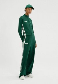 Chándal deportivo verde que incluye una chaqueta con cremallera y pantalones de pierna ancha, acentuado con rayas y logotipos blancos. Combinado con una gorra a juego.