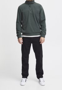 Sudadera de forro polar verde con media cremallera, puños y dobladillo acanalados. Lleva puesta sobre una camisa blanca, combinada con pantalones negros y zapatillas blancas.