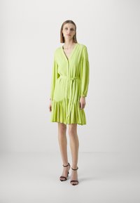 Lime green dress made of lightweight fabric, featuring a V-neck, button closure, long sleeves, tie belt, and ruffled hem. Black sandals.