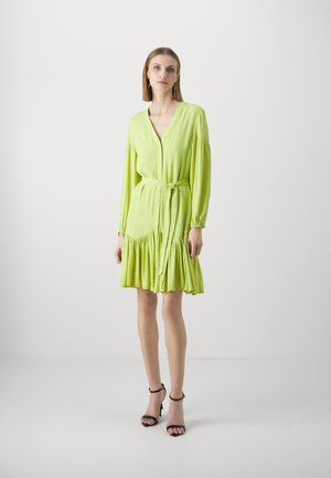 Lime green dress made of lightweight fabric, featuring a V-neck, button closure, long sleeves, tie belt, and ruffled hem. Black sandals.