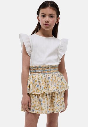 Jeune fille portant un haut blanc sans manches à volants et une jupe fleurie à volants avec une taille smockée, debout devant un fond uni.