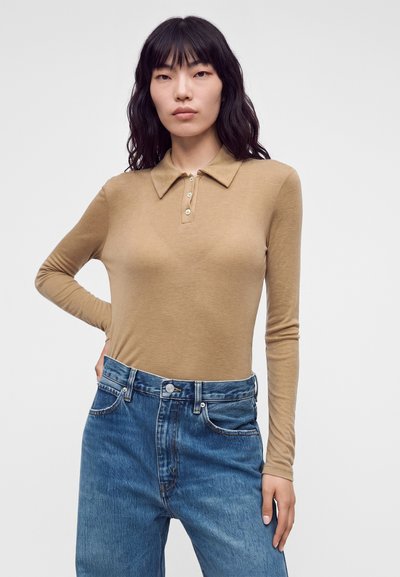 Femme aux cheveux bruns ondulés porte un polo beige à manches longues rentré dans un jean bleu taille haute, debout avec une main sur la hanche.