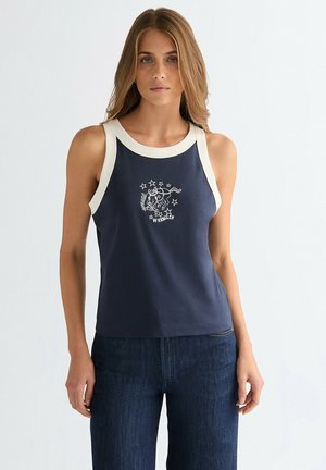 Kvinde iført navyblå tanktop med hvid kant og stjerne- og hestegrafik, kombineret med mørkeblå jeans, stående mod en lys baggrund.