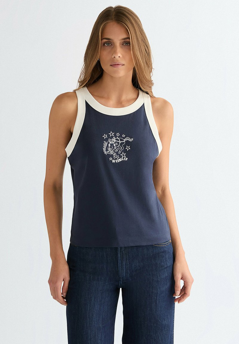 Vrouw draagt een marineblauw tanktop met witte bies en een ster- en paardenmotief, gecombineerd met donkerblauwe jeans, staand tegen een lichte achtergrond.