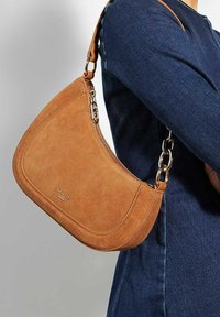 Suède handtas in tan met een gebogen vorm. Voorzien van een ritssluiting en een kettingband. Glad textuur met subtiele stikdetails.