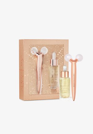Ensemble de trois outils de soin en cristal : rouleaux en jade rose et or rose, deux pipettes en verre avec des sérums scintillants, dans un emballage en boîte pailletée.