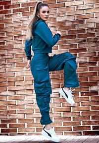 Felpa corta teal con dettagli neri, abbinata a pantaloni cargo teal con molte tasche e sneaker basse bianche con dettagli neri.