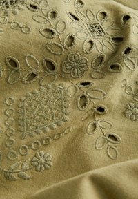 Tissu vert olive présentant une broderie complexe avec des motifs floraux et géométriques. Texturé avec des détails ajourés et des points surélevés.