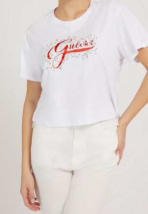Hvid crop top med rød "Guess"-logo og små sølvdekorationer, båret med hvide bukser på en stående person.