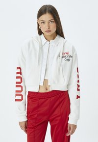 Sudadera blanca cortada con texto rojo en la manga izquierda y detalle de lazo bordado en rosa. Combinada con pantalones deportivos rojos, textura suave y diseño casual.