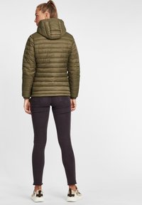 Olivna puffer jakna s horizontalnim šivom in kapuco, v kombinaciji s temno ozkimi kavbojkami in bež supergami z black akcentom.