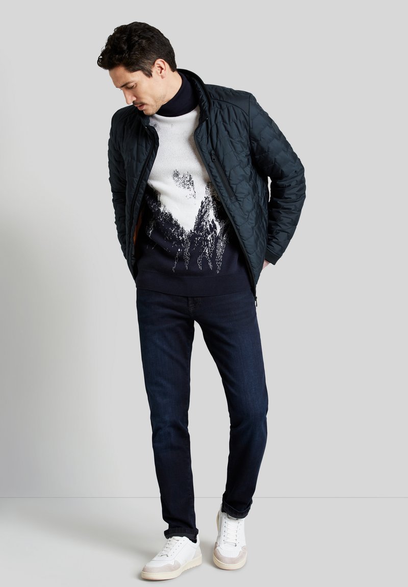 Un homme portant une veste matelassée noire, un pull graphique noir et blanc, un jean foncé et des baskets blanches, sur un fond gris uni.