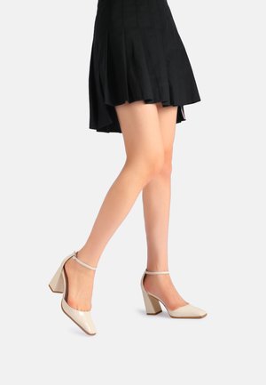Scarpe con tacco alto in pelle verniciata beige, con punta quadrata e cinturino alla caviglia. Presentano un tacco a blocco e una finitura liscia e lucida.