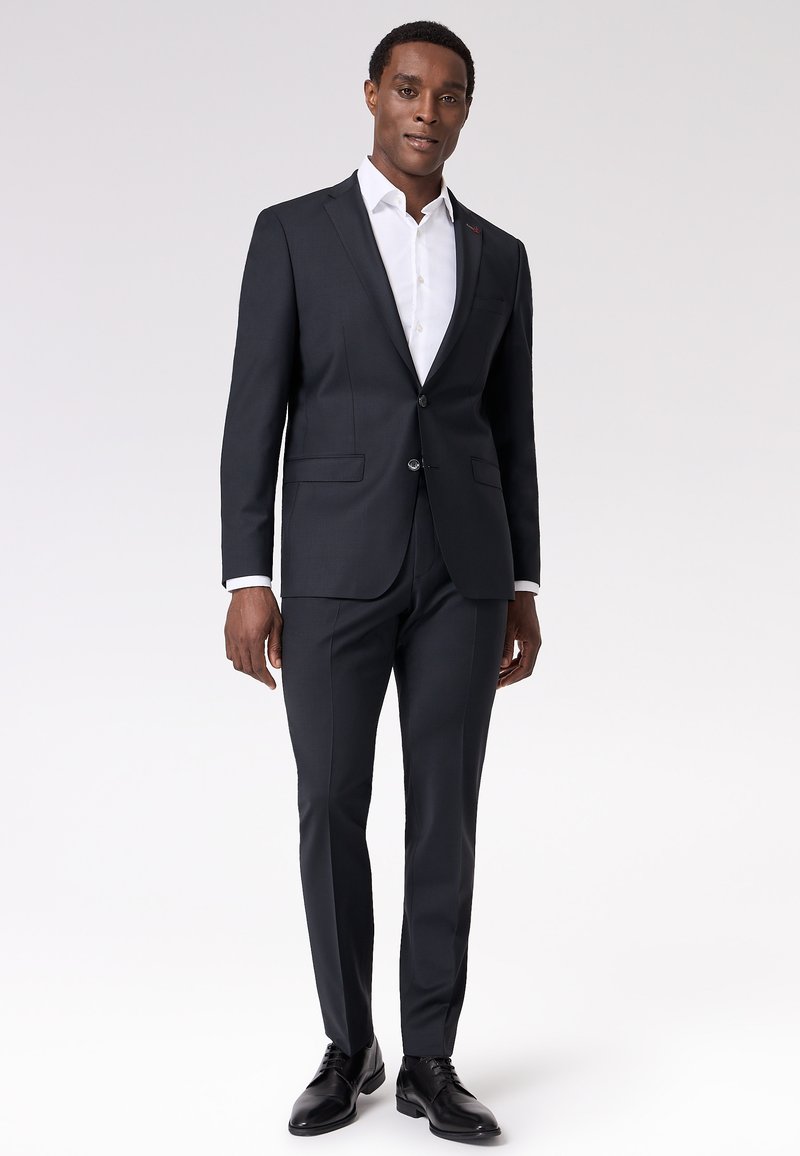 Completo su misura grigio scuro con giacca a un bottone, pantaloni slim-fit e camicia bianca con colletto. Tessuto liscio, design minimalista.