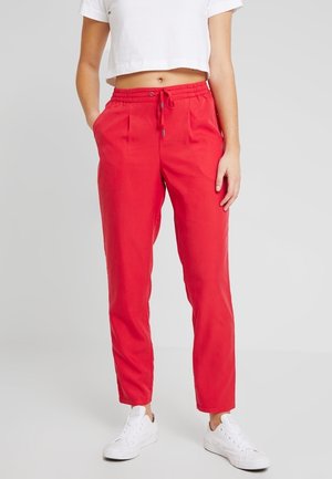 Frau trägt leuchtend rote Jogginghosen mit Kordelzug, weißes kurzgeschnittenes T-Shirt und weiße Turnschuhe, steht vor einem einfarbigen Hintergrund.